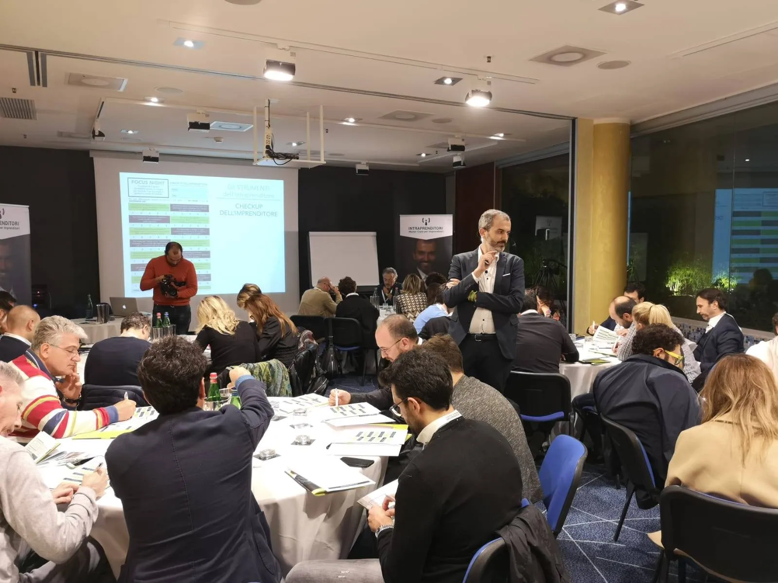 Sala di un workshop Intraprenditori con imprenditori al lavoro sui materiali e Andrea Lagravinese fra i tavoli